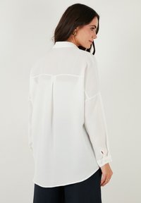 White long-sleeve shirt with a textured fabric, a collared neckline, and buttoned cuffs. Features a back yoke and pleat detail.