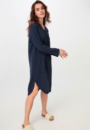 Marineblaue, langärmelige Kleider aus strukturiertem Stoff, mit einer Knopfleiste und seitlichen Schlitzen für Bewegungsfreiheit; kombiniert mit beigen Slip-On-Sandalen.