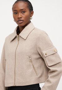 Kragen beige ullblandad jacka med avskuren längd, framzipning, två fickor med dragkedja och en knappficka på vänster ärm.