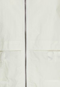 Béžová tkanina se středním kovovým zipem a prošívanými kapsami po obou stranách, detailní záběr na část oděvu.