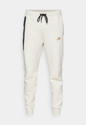 Pantaloni sportivi Nike color crema con gambe affusolate, cintura elastica, lacci neri e tasche laterali con zip. Logo dorato sulla coscia sinistra.