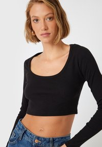 Top crop nero a maniche lunghe con scollatura a barca, realizzato in tessuto a costine, abbinato a jeans in denim blu a vita alta con dettaglio dei bottoni.
