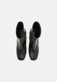 Bottines en cuir noir avec une texture lisse, bout pointu, détail de fermeture éclair sur le côté et étiquette de marque visible à l'intérieur.