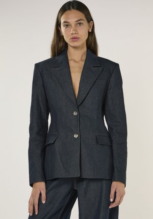 Donna che indossa un blazer strutturato in denim blu scuro con due bottoni d'argento e pantaloni in denim abbinati, in piedi contro uno sfondo semplice.