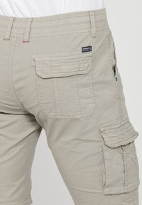 Beiga cargoshorts med ficklock på baksidan och sidan, med synliga sömmar och en liten tyglogotagg nära midjan.