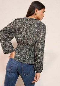 Blusa a maniche lunghe in tessuto nero con macchie bianche, caratterizzata da una vita arricciata e polsini elastici. Abbinata a jeans in denim blu.