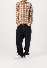 Man in een bruin en wit geruite lange mouwen shirt, losse zwarte broek, witte sneakers, staande met zijn handen in zijn zakken tegen een effen achtergrond.