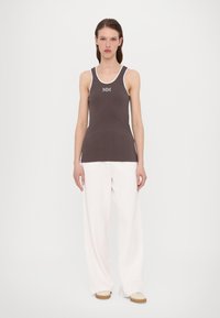 Braun geripptes Tanktop mit weißen Besätzen und "MM"-Logo, kombiniert mit lockeren weißen Hosen. Das Modell steht vor einem schlichten weißen Hintergrund.