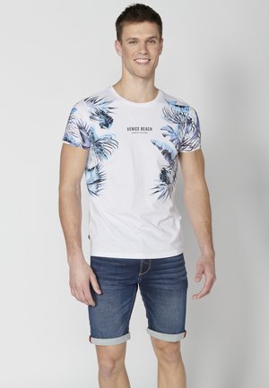 White short-sleeve T-shirt featuring blue tropical leaf prints on shoulders, with "VENICE BEACH" text. Worn with denim shorts, rolled cuffs.