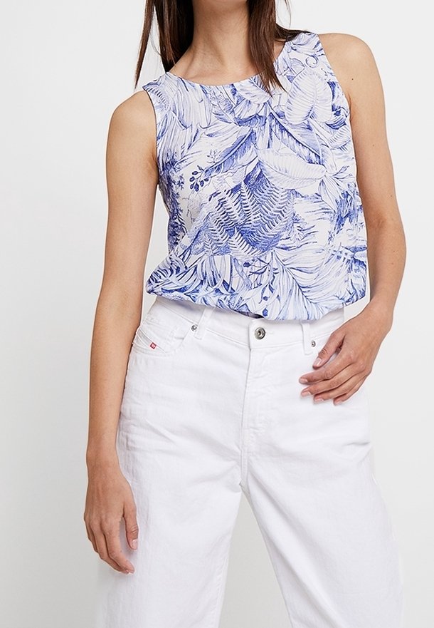 Femme portant un haut blanc sans manches à imprimé feuilles bleues et un pantalon blanc taille haute, posant avec une main sur la hanche et l'autre détendue.