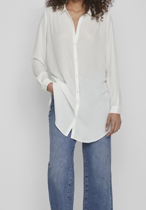 Woman with curly hair wearing a loose white button-up shirt and blue jeans, standing with one hand in pocket against plain background.