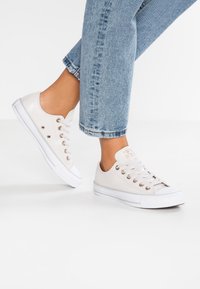 Canvas sneakers i off-white med metallöglor, vita gummisulor och en kontrasterande tåkappa; stylade med mönstrade ljusblå jeans.