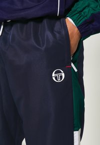 Marinové sportovní kalhoty s lesklou texturou, s bílým logem a kontrastními zelenými bočními panely s detaily červeného prošívání.