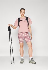 Rosa kortermet skjorte med lomme, mønstrede rosa og lilla shorts, grå tursko, svart ryggsekk og fjellstaver.
