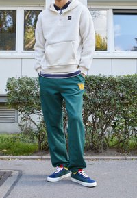 Fleecehoodie i krämfärg, gröna sweatpants med gult "W"-patch, och flerfärgade sneakers. Mot en bakgrund av buskar och asfalt.