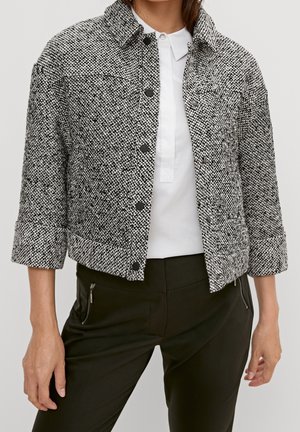 Kvinde iført en sort og hvid tekstureret jakke med knapper, hvid skjorte med krave og sorte bukser med lynlåslommer.