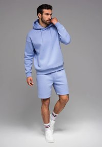 Felpa leggera blu con cappuccio e cordini, abbinata a pantaloni corti. Presenta una vestibilità comoda, materiale morbido, polsini a coste e dettagli con logo sulla manica.