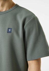 Grijze, gestructureerde T-shirt met korte mouwen en een vierkante logo-patch op de borst. Heeft een ronde halslijn en een losse pasvorm.