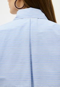 Camicia blu a righe con linee verticali di diverse larghezze. Presenta un colletto classico e un dettaglio di cucitura sul retro. Tessuto di consistenza liscia.