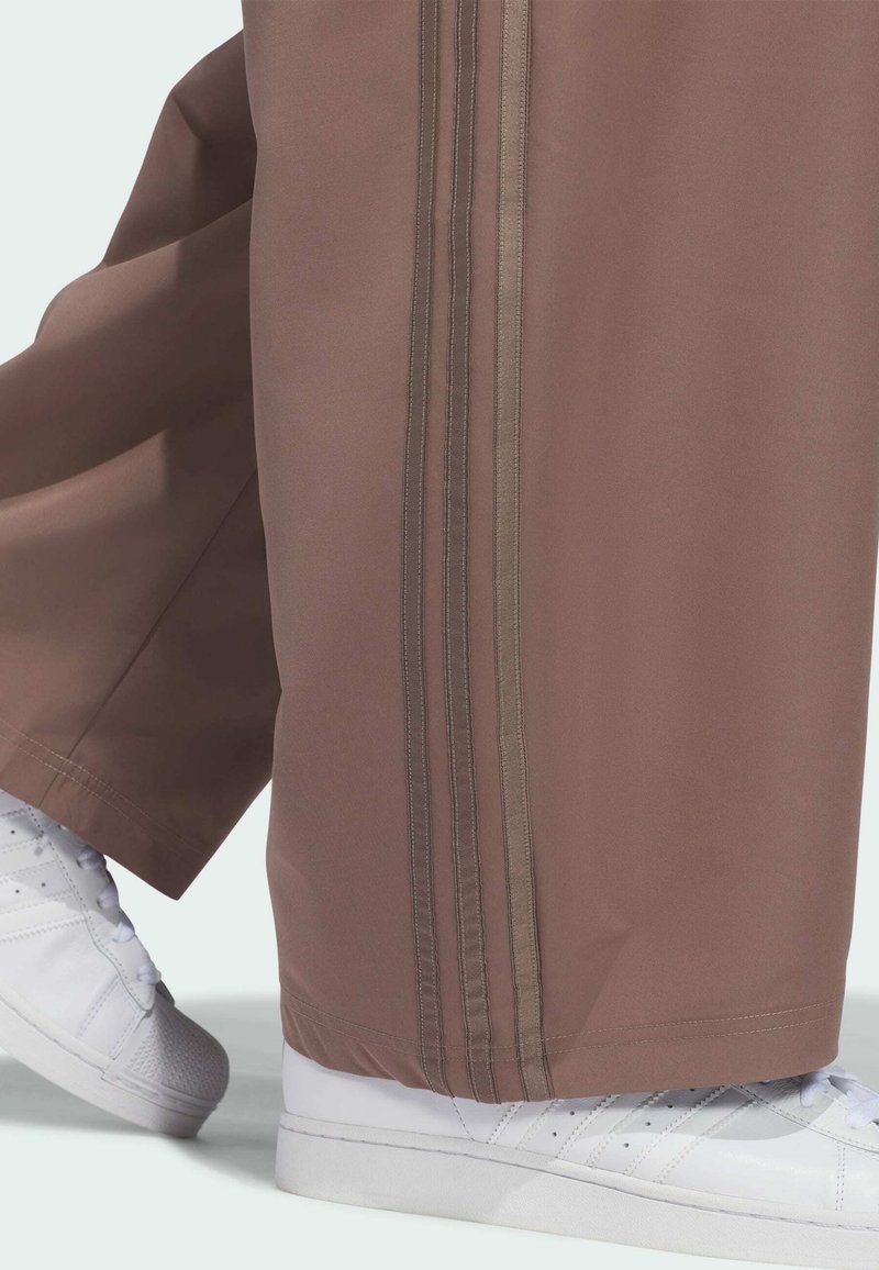 Brown wide-leg pants with a double vertical stripe detail worn over white sneakers with laces on a light background.