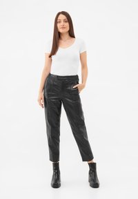 Pantalon en cuir noir ajusté avec des poches latérales, associé à un t-shirt blanc à col en V près du corps et des bottines noires, sur un fond uni.