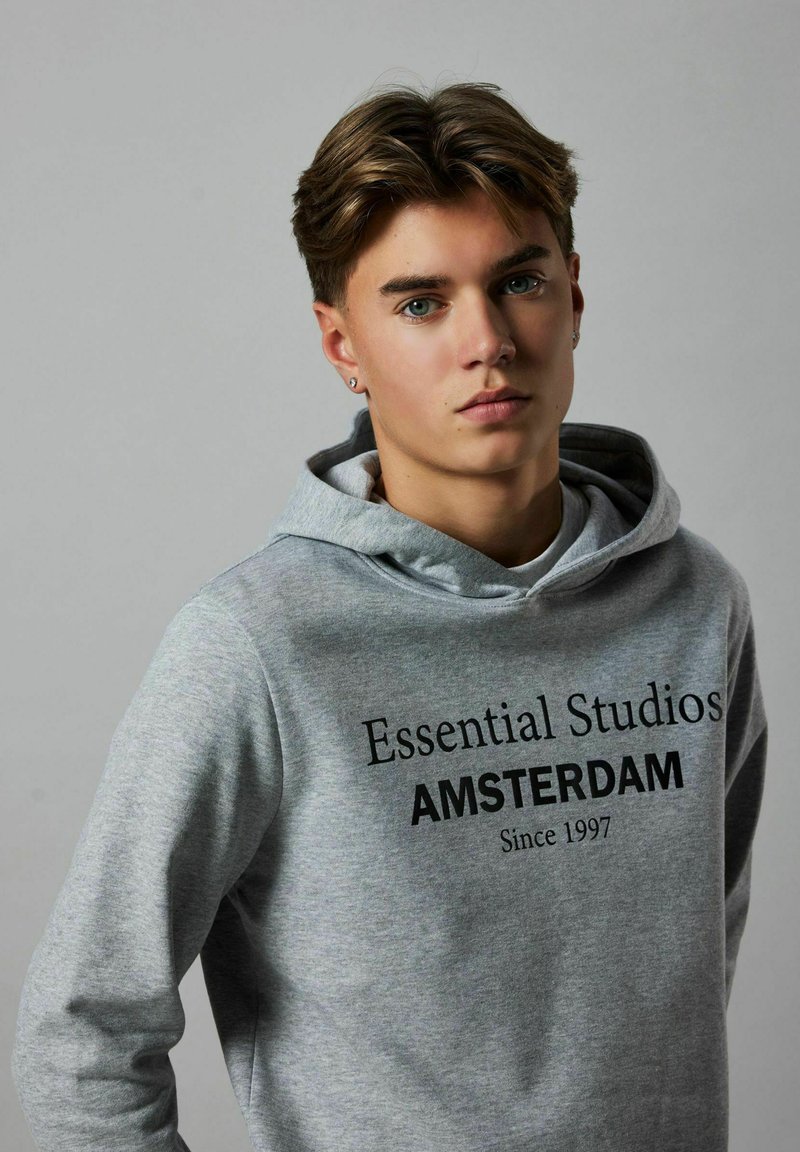 Mlađi muškarac s svijetlosmeškom kosom nosi sivi duksericu s natpisom "Essential Studios AMSTERDAM Since 1997" na jednostavnoj pozadini.
