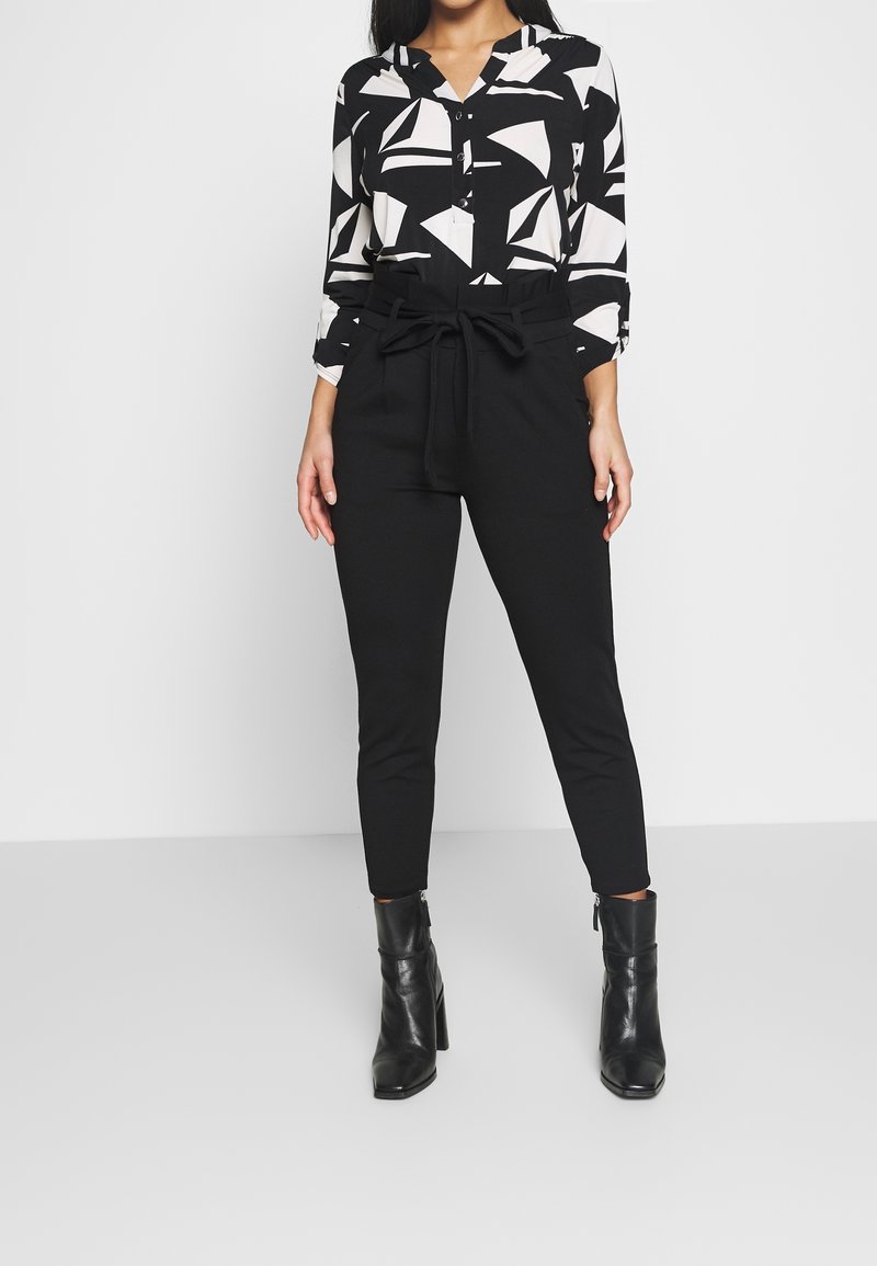 Femme portant un chemisier à motifs géométriques noir et blanc, un pantalon noir taille haute avec ceinture nouée et des bottines noires, debout sur un sol gris.