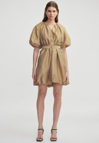 Tan jurk met pofmouwen, een textuurafwerking, een taille die wordt aangesnoerd door een riem en een zacht geplooide rok. Draag het met zwarte sandalen met bandjes.