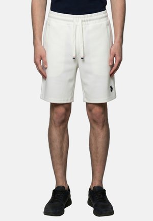 Man draagt witte short met trekkoord, zijzakken en een zwart logo op het linkerbeen, gecombineerd met zwarte sneakers en een donker topje.