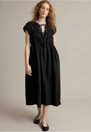 Femme aux cheveux bouclés portant une robe midi noire ample avec des liens à l'encolure et des chaussures plates noires, debout sur un fond uni.