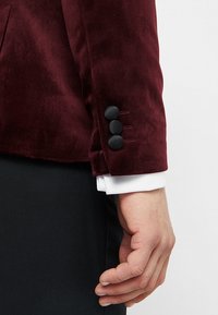 Maronowy aksamitny żakiet z rękawem wykończonym czterema czarnymi guzikami, charakteryzujący się gładką teksturą i dopasowanym krojem, w połączeniu z mankietem białej koszuli.