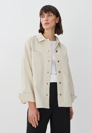 Beige button-up shirt with a collar, large front pockets, and rolled-up sleeves. Made of a textured fabric, displayed over a white top.