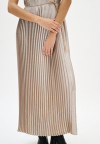 Plisowana sukienka maxi w jasnym neutralnym kolorze, z talią podkreśloną paskiem oraz fakturowaną tkaniną. Dopełniona czarnymi botkami do kostki.