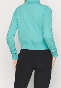 Felpa azzurra chiara, cropped, con colletto alto, polsini e orlo a coste; indossata con pantaloni neri dotati di tasca posteriore.