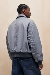 Grå bomberjakke med en glat, skinnende tekstur, ribbede manchetter og kant. Har en bred krave og afslappet pasform, parret med mørk denim jeans.