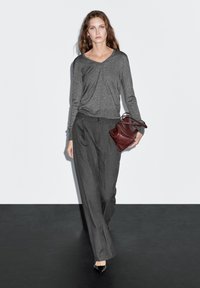 Gray long-sleeve top with a V-neck, paired with high-waisted pinstriped trousers. Model holds a burgundy leather handbag. Black pointed-toe shoes.