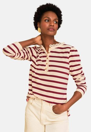 Vrouw met kort krullend haar, gouden oorbellen, een crème- en bordeaux gestreept shirt met lange mouwen en crèmekleurige broek, poserend met één hand in de nek.