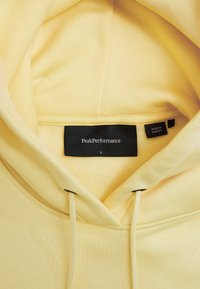 Gelbe Kapuzenjacke mit weicher Textur; enthält ein schwarzes Etikett mit der Aufschrift "PeakPerformance", Größe S, und ein kleineres Etikett, das "Hergestellt in der Türkei" anzeigt.
