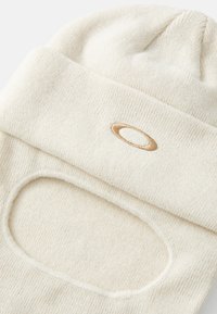 Balaclava tricot beige avec une ouverture circulaire pour le visage, dotée d'un logo cousu et d'un tissu texturé pour la chaleur.