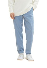 Osoba w jasnoniebieskich spodniach i białych trampkach, z jedną ręką w kieszeni i częściowo widocznym białym swetrem.