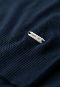 Marinblå stickad tröja med ribbad textur, med en silvermetallmärkning präglad med "QUINTESSENCE" sydd längst ner.