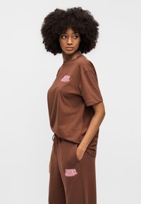 Καφέ βαμβακερό t-shirt με άνετη εφαρμογή και κοντά μανίκια, με ροζ κεντημένο λογότυπο Nike στο στήθος. Στυλιζαρισμ ένο με ταιριαστό παντελόνι.