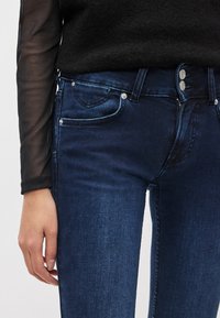 Vaqueros pitillo azul oscuro con textura suave, con dos bolsillos delanteros, botones plateados y cremallera. Llevados con una blusa negra transparente de manga larga.