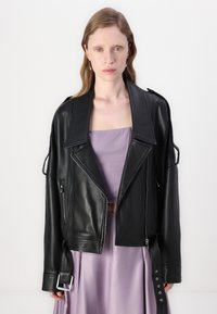 Jaqueta de motoqueiro cropped em couro preto com lapelas largas, bolsos com fecho éclair e detalhes de tira ajustável. Usada sobre uma blusa lavanda.