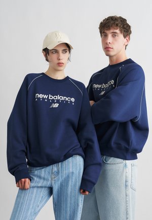 Dwóch młodych dorosłych ubranych w granatowe bluzy New Balance Athletics i jasnoniebieskie dżinsy stoi na tle jednolitego jasnego tła.