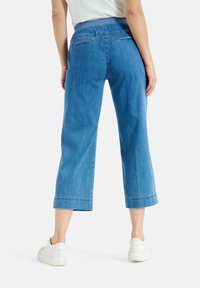Pantalons en denim bleu, cropped, avec une jambe large, présentant une taille lisse, des poches arrière et un ourlet soigné. Portés avec des baskets blanches.