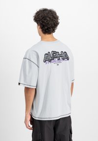 Šedé bavlněné tričko s oversized střihem, krátkými rukávy, na zadní straně černý a fialový grafický text "ALPHA STUDIO EDITION". Kontrastní švy.