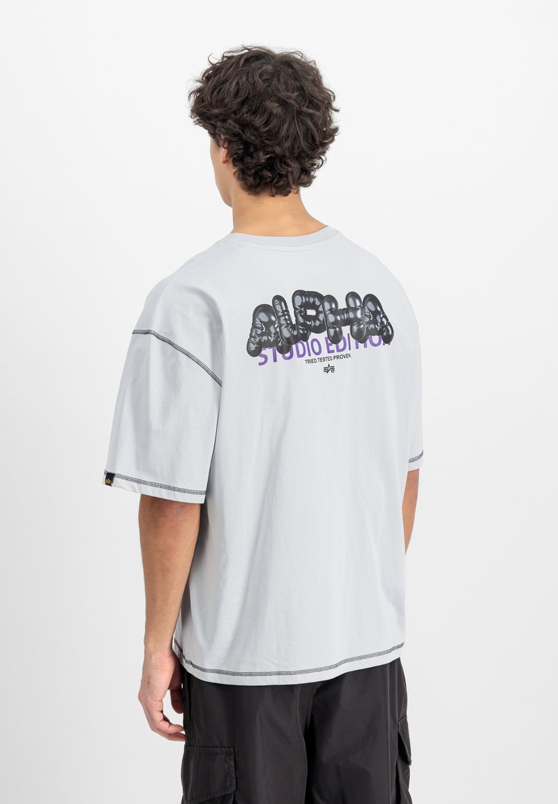 Šedé bavlněné tričko s oversized střihem, krátkými rukávy, na zadní straně černý a fialový grafický text "ALPHA STUDIO EDITION". Kontrastní švy.