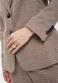 Kariertes Blazer in Beige, Braun und Schwarz. Verfügt über ein tailliertes Design, schwarze Knöpfe und strukturierte Ärmel. Hand mit Ringen auf der Taille positioniert.