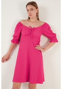 Fuchsia dress with off-shoulder design, puff sleeves, and a front tie detail. Made of textured fabric, ending just above the knees.